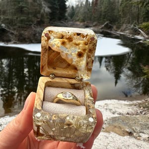 Pode incluir: Um anel de ouro com uma pedra redonda transparente inserido numa caixa quadrada de resina e madeira. A caixa apresenta flores secas incrustadas e texturas naturais, com um encaixe de tecido claro que segura o anel no centro.