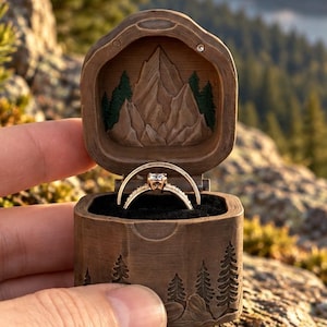 May include: A brown wooden ring box featuring a carved mountain and forest landscape on the interior lid and exterior. The box contains two gold rings with small stones, set against a black velvet interior lining, held in a horizontal position.