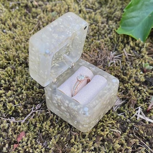 Op de afbeelding: Een open, heldere hars ringdoos met een gouden verlovingsring erin, geplaatst op een bedje van lichtroze stof. De doos staat op een bedje van groen mos, met groene bladeren op de achtergrond.
