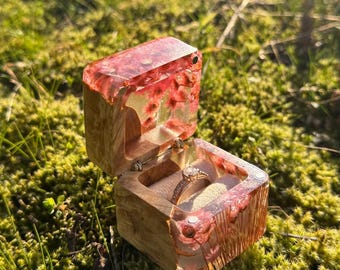 May include: Open wooden ring box with a clear resin top and sides, embedded with red flowers. The box is open, revealing a gold ring with a diamond. The box sits on a bed of green moss.
