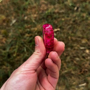 May include: A ring made of translucent pink resin with embedded gold-colored metallic flakes. The band has a smooth, rounded exterior surface and a glossy finish, showcasing the suspended glitter particles throughout the material.