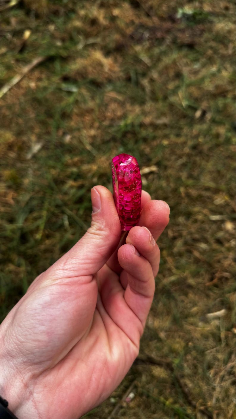 May include: A ring made of translucent pink resin with embedded gold-colored metallic flakes. The band has a smooth, rounded exterior surface and a glossy finish, showcasing the suspended glitter particles throughout the material.
