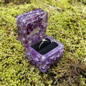 May include: A purple resin ring box with embedded flowers, holding a diamond ring. The box is open, revealing a black velvet interior. The box is set against a backdrop of green moss and grass.