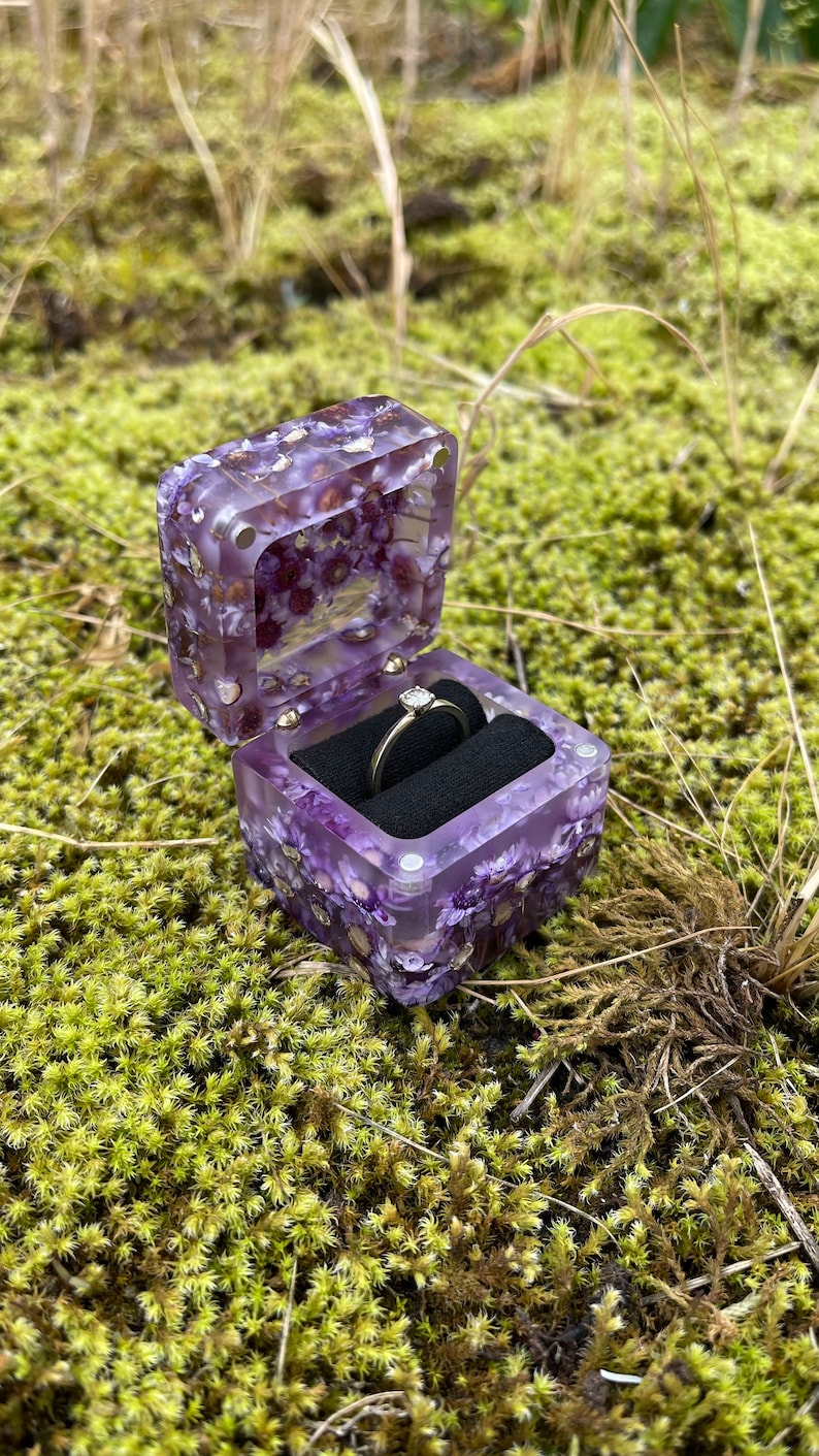 May include: A purple resin ring box with embedded flowers, holding a diamond ring. The box is open, revealing a black velvet interior. The box is set against a backdrop of green moss and grass.