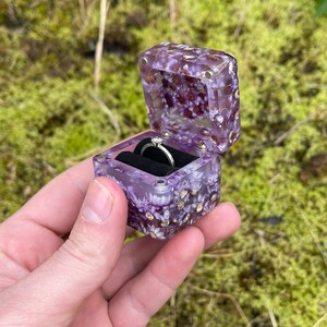 May include: A square, purple resin ring box with embedded dried flowers and gold flakes. The box is open, revealing a silver ring with a small diamond. The box is held against a blurred green background.