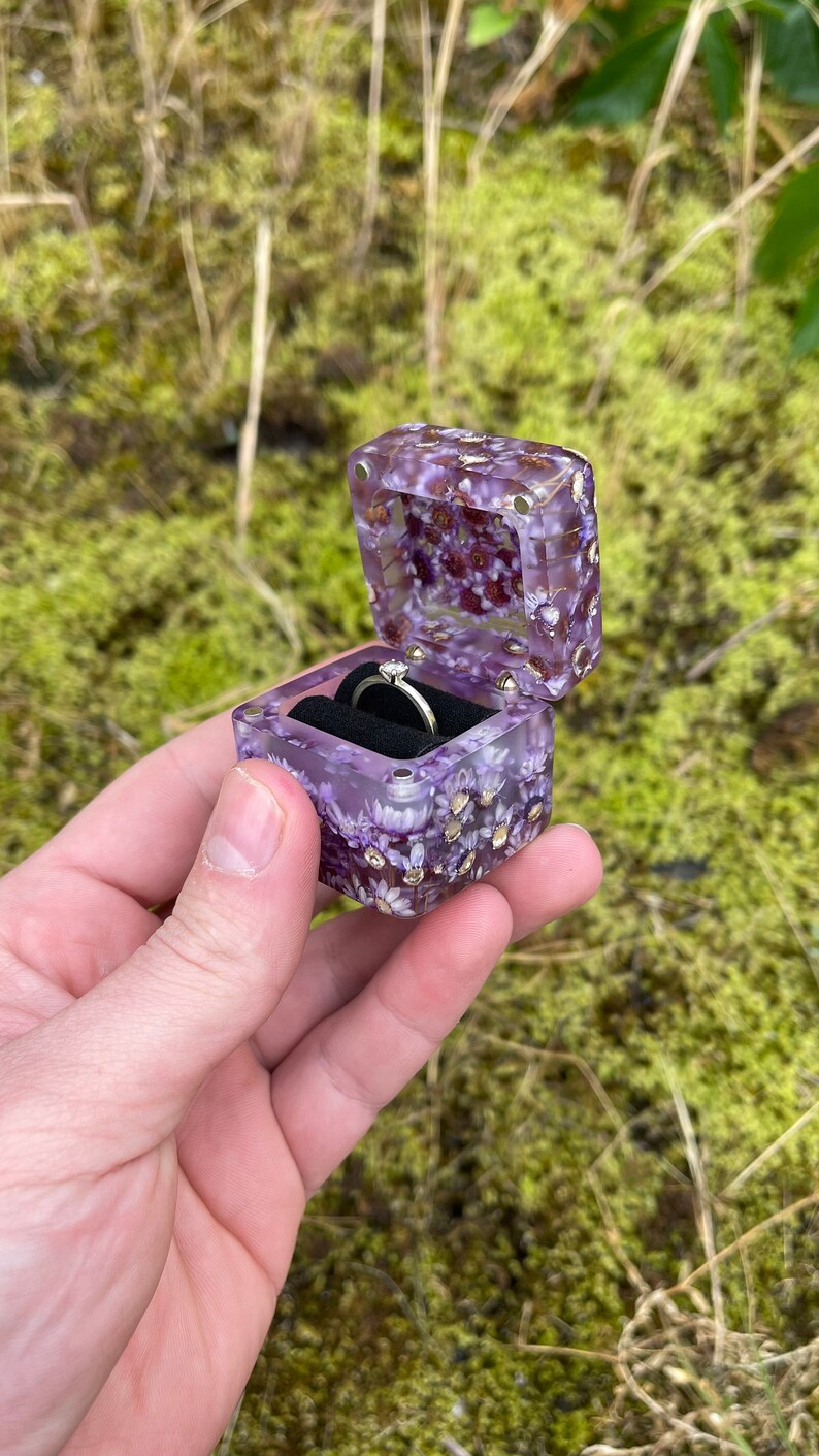 May include: A square, purple resin ring box with embedded dried flowers and gold flakes. The box is open, revealing a silver ring with a small diamond. The box is held against a blurred green background.