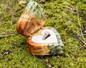 May include: A heart-shaped ring box crafted from natural wood and translucent resin containing embedded dried white flowers. The open box features a light-colored velvet interior lining holding a silver-toned ring with a single clear stone.