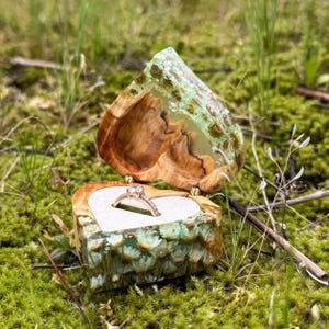 Puede incluir: Un joyero en forma de corazón de madera con un exterior de resina verde transparente y un interior claro. La caja está abierta, revelando un anillo de oro con un diamante. La caja está colocada en un lecho de musgo verde.