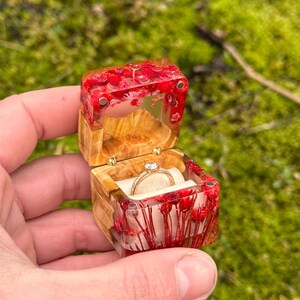 Può includere: Una piccola scatola di legno per anelli con un coperchio trasparente riempito di fiori pressati rossi e un anello d'oro con un solo diamante.