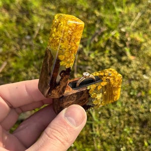 Puede incluir: Un joyero artesanal de madera y resina, con un anillo de oro con un diamante. La caja es rectangular con tapa con bisagras, y la resina es amarilla con elementos florales incrustados. La caja está abierta, revelando el anillo.