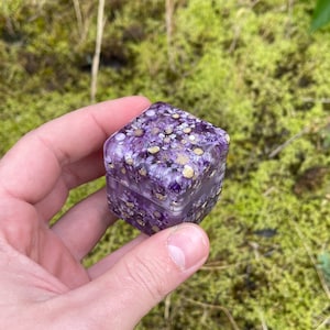 May include: A small, square box with a hinged lid, filled with purple flowers and gold flecks. The box is held in a hand against a backdrop of green moss and grass. The box is approximately 2 inches wide and 2 inches tall.