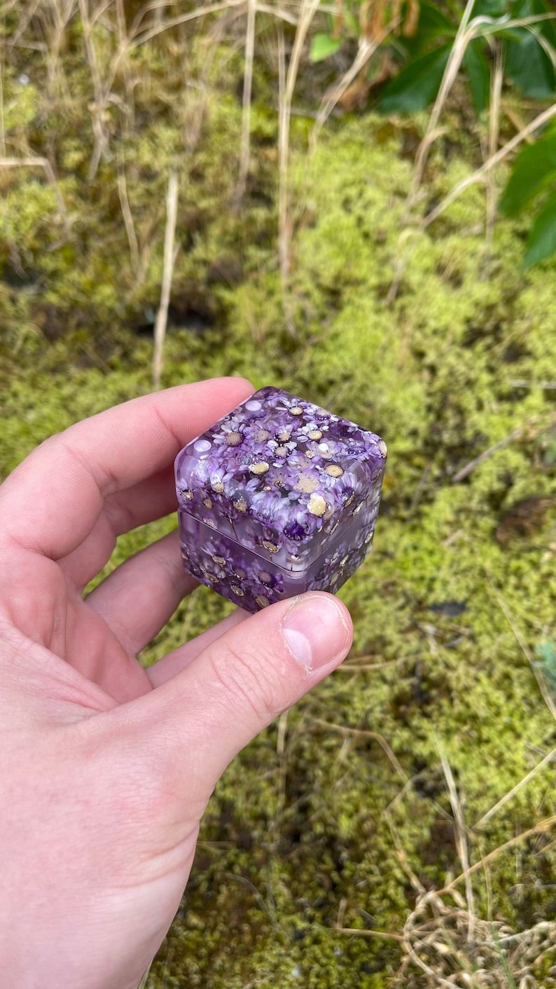 May include: A small, square box with a hinged lid, filled with purple flowers and gold flecks. The box is held in a hand against a backdrop of green moss and grass. The box is approximately 2 inches wide and 2 inches tall.
