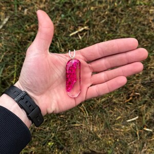 May include: A clear, rectangular pendant filled with vibrant pink flowers. The pendant has a silver bail and is held in an open hand against a backdrop of green grass. The watch on the wrist is black.