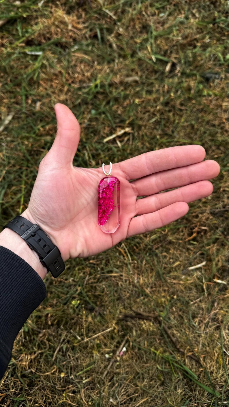 May include: A clear, rectangular pendant filled with vibrant pink flowers. The pendant has a silver bail and is held in an open hand against a backdrop of green grass. The watch on the wrist is black.