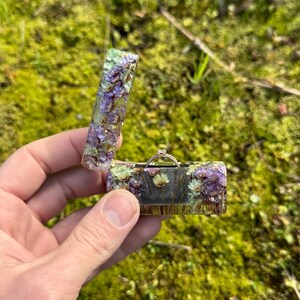 Peut inclure: Une petite boîte à bague rectangulaire en résine transparente avec des fleurs séchées incrustées dans des tons de violet, vert et blanc. La boîte est ouverte, révélant une bague en or avec un seul diamant.