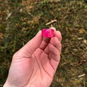 May include: A vibrant pink resin ring with a rectangular shape is held in a hand. The ring has a marbled appearance with lighter pink and white swirls. The background is blurred green grass.