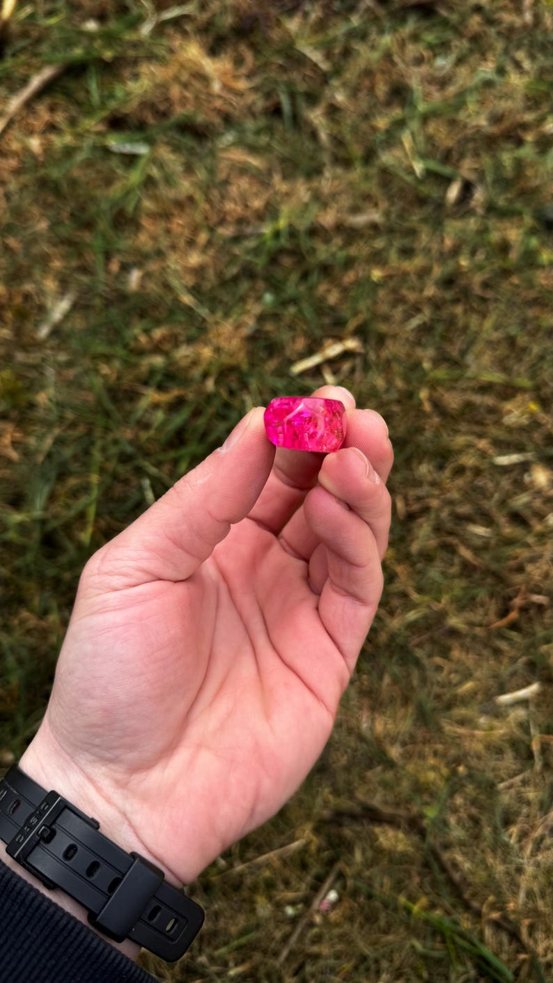 May include: A vibrant pink resin ring with a rectangular shape is held in a hand. The ring has a marbled appearance with lighter pink and white swirls. The background is blurred green grass.