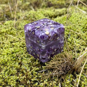May include: A square, clear resin box filled with purple flowers and gold flecks. The box is sitting on a bed of green moss and is surrounded by dried grass.