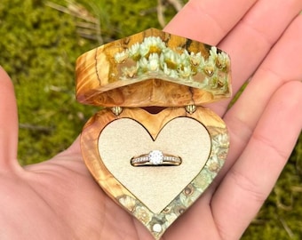 May include: A heart-shaped wooden ring box with a hinged lid, open to reveal a diamond engagement ring. The box is made of light-colored wood with a floral resin design. The ring has a large, round diamond.