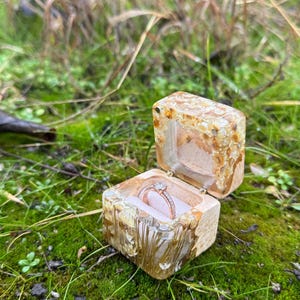 May include: A small, wooden ring box with a clear lid, filled with gold and white resin. The box is open and a diamond ring is inside.