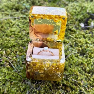 May include: A small wooden ring box with a clear lid, filled with dried flowers and a gold ring with a single diamond. The box is sitting on a bed of green moss.