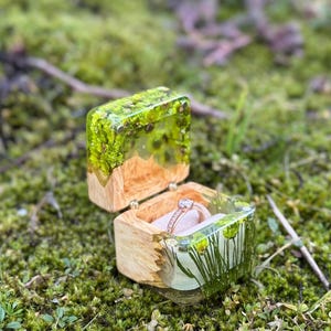 Puede incluir: Una pequeña caja de madera para anillos con una tapa transparente llena de plantas y flores verdes preservadas. Un anillo de oro con un solo diamante está dentro de la caja.
