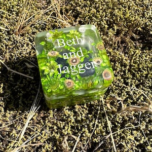 May include: A clear, square resin paperweight with the names "Beth and Jagger" in white lettering. The paperweight is filled with green plants and small, brown, flower-like accents. The background is moss and dry grass.