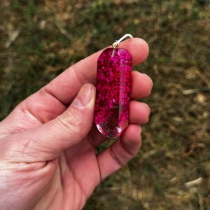 May include: A clear resin pendant shaped like a faceted crystal, filled with vibrant magenta dried flowers. The pendant features a small silver-toned metal bail at the top for attachment, showcasing a smooth, polished finish and a geometric form.