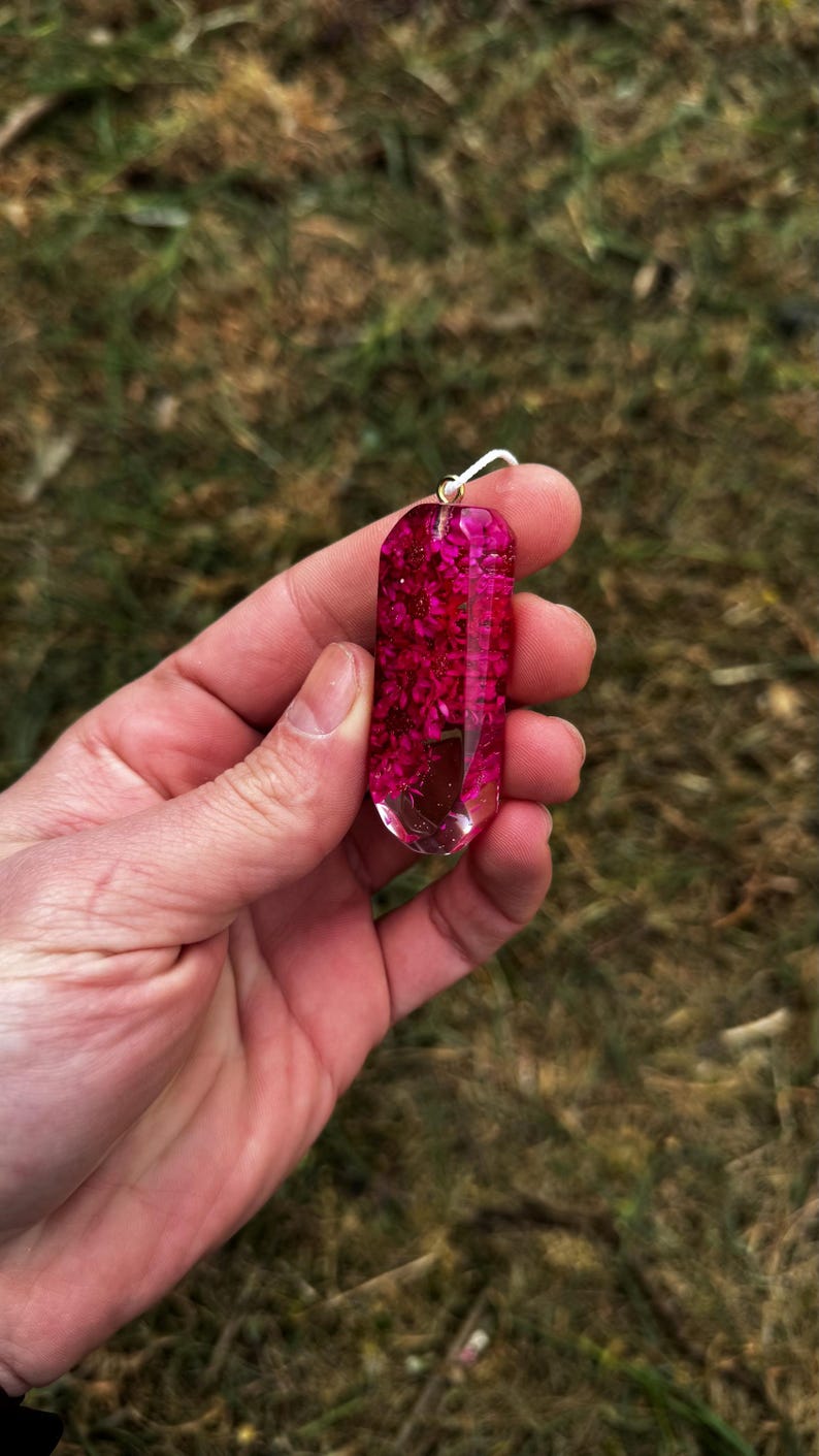 May include: A clear resin pendant shaped like a faceted crystal, filled with vibrant magenta dried flowers. The pendant features a small silver-toned metal bail at the top for attachment, showcasing a smooth, polished finish and a geometric form.