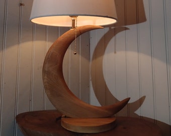 May include: A white lampshade with a crescent moon shaped wooden base. The lamp is on a wooden table.