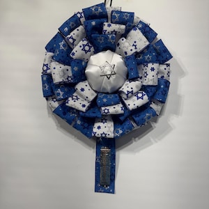 May include: A decorative wreath featuring blue and white ribbons with silver Star of David patterns. A silver satin kippah is in the center, and a matching ribbon with a silver mezuzah case hangs below. The wreath is designed for Jewish celebrations.