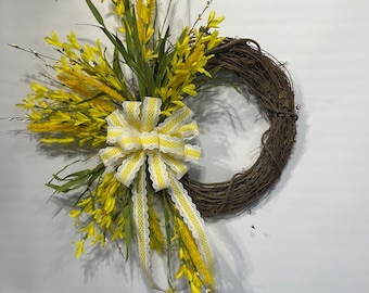 May include: A yellow and white floral wreath with a yellow and white polka dot bow. The wreath is made of grapevine and is decorated with yellow flowers and green leaves.