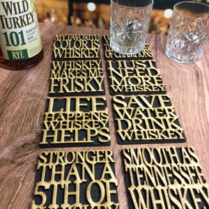 May include: Set of eight black and gold wooden coasters with humorous sayings about whiskey. The sayings include "My favorite color is whiskey", "Whiskey makes me frisky", "Life happens whiskey helps", "Stronger than a 5th of whiskey", "Whiskey of champions", "I just need more whiskey", "Save water drink whiskey", and "Smooth as Tennessee whiskey".