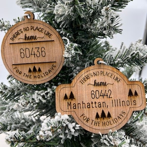 May include: Two wooden Christmas ornaments with the text "There's no place like home" and a zip code. The top ornament has the zip code "60436" and the bottom ornament has the zip code "60442" and the text "Manhattan, Illinois". Both ornaments have a small tree design and the text "For the Holidays".