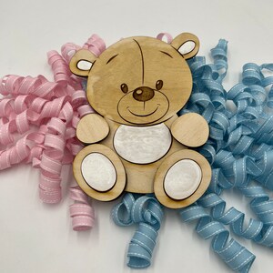 May include: A wooden bear cutout with a smiling face, surrounded by pink and blue curled ribbon.