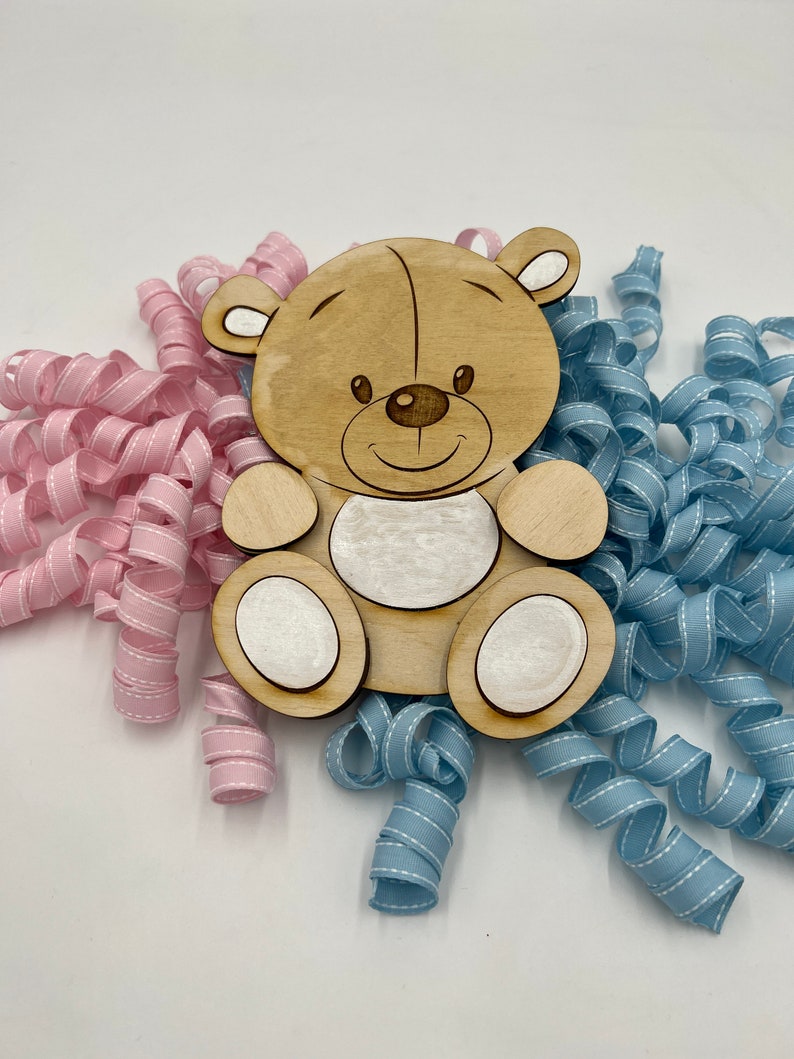 May include: A wooden bear cutout with a smiling face, surrounded by pink and blue curled ribbon.