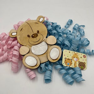 May include: A wooden cutout of a brown teddy bear with a white face and white paws. The bear is surrounded by blue and pink curled ribbon. A small card with a giraffe, tiger, and bear is in the lower right corner.