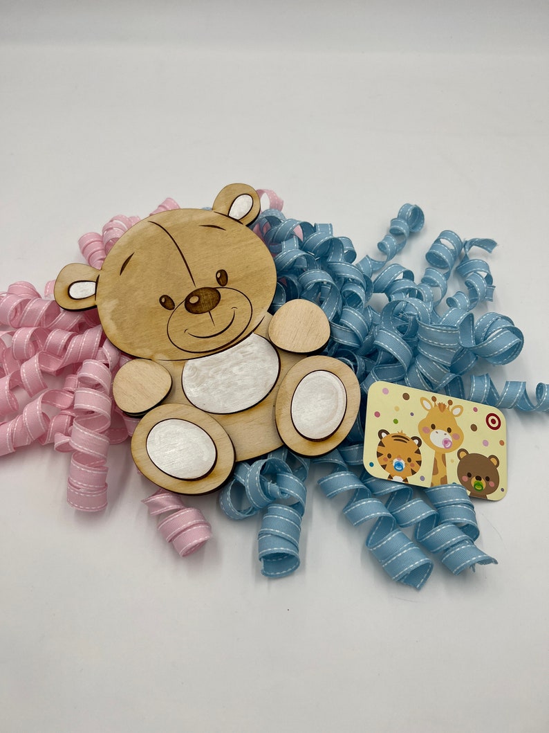 May include: A wooden cutout of a brown teddy bear with a white face and white paws. The bear is surrounded by blue and pink curled ribbon. A small card with a giraffe, tiger, and bear is in the lower right corner.
