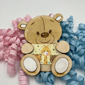 May include: Wooden bear figurine with a light brown finish. The bear is holding a card with a giraffe, tiger, and bear illustration. The card has a yellow background with brown polka dots. The bear is sitting on a pink and blue ribbon.