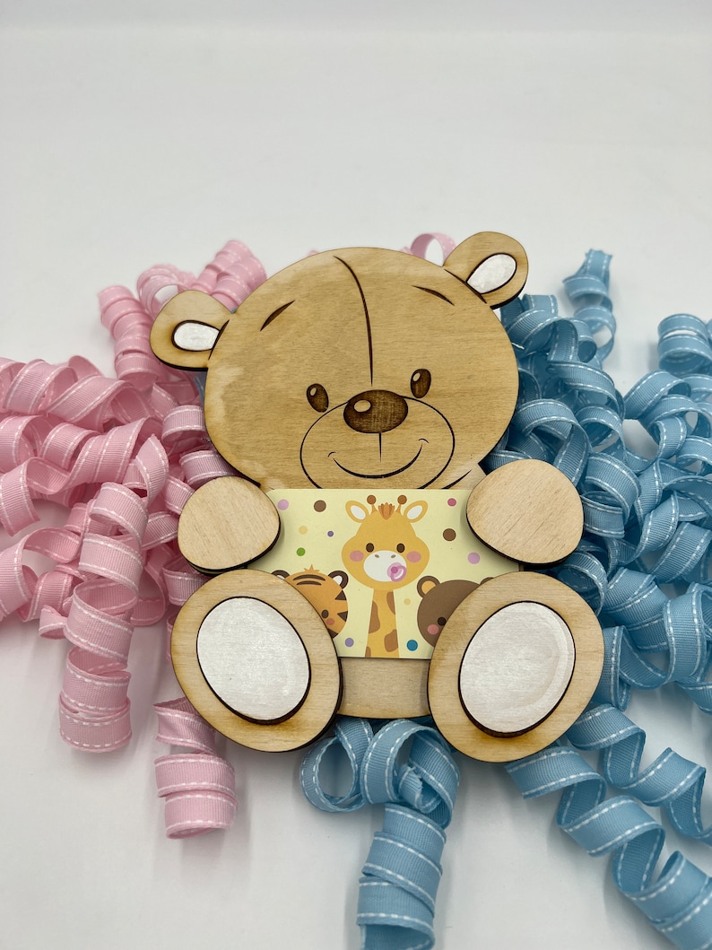 May include: Wooden bear figurine with a light brown finish. The bear is holding a card with a giraffe, tiger, and bear illustration. The card has a yellow background with brown polka dots. The bear is sitting on a pink and blue ribbon.