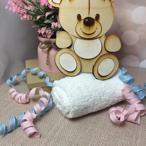 May include: A wooden cutout of a smiling teddy bear sitting on a white towel. The bear is light brown with a white belly and a brown nose. The towel is rolled up and there is a pink and blue ribbon on the table.