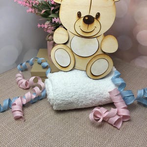 May include: A wooden cutout of a teddy bear with a smiling face. The bear is sitting on a white towel with pink and blue ribbon.