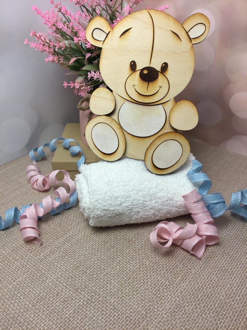 May include: A wooden cutout of a teddy bear with a smiling face. The bear is sitting on a white towel with pink and blue ribbon.