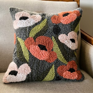 Puede incluir: Una almohada gris con un diseño floral. La almohada presenta flores rosas y naranjas con hojas verdes. Las flores están bordadas en un estilo elevado y texturizado.
