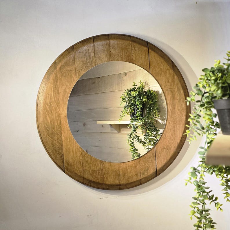 May include: Circular wooden mirror with a natural wood grain finish. The mirror is set in a thick, circular frame. The reflection shows a small shelf with a potted plant. The mirror is mounted on a white wall.
