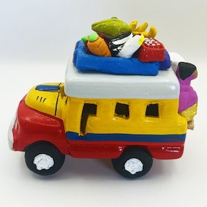 May include: A yellow and red toy bus with a blue roof and a white top. The bus is carrying a variety of colorful fruits and vegetables, including a green avocado, a carrot, and a banana. The bus is a traditional Mexican folk art piece.