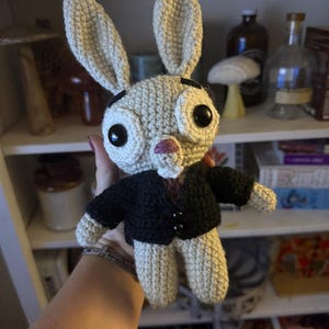 May include: A handmade crochet bunny doll with large black eyes, long ears, and a small pink nose. The bunny wears a black jacket with two buttons. The doll is beige and has a whimsical, cartoonish design.