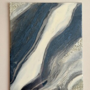 May include: A rectangular wall art piece featuring a glossy resin finish with swirling patterns of deep navy blue, white, and gray. The composition includes diagonal bands of color accented by textured, shimmering silver crushed glass elements.