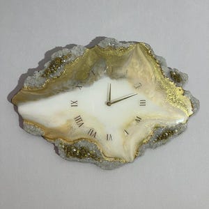May include: An irregular, geode-inspired wall clock featuring a smooth white and cream center with gold accents. The clock face displays gold Roman numerals and thin gold hands. The outer edge has a textured, crystalline gray and gold border.