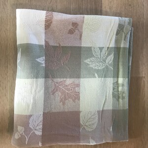 May include: Folded rectangular tablecloth with a checkered pattern in shades of cream, green, and brown. The design features embossed leaf motifs. The tablecloth is made of a soft fabric.